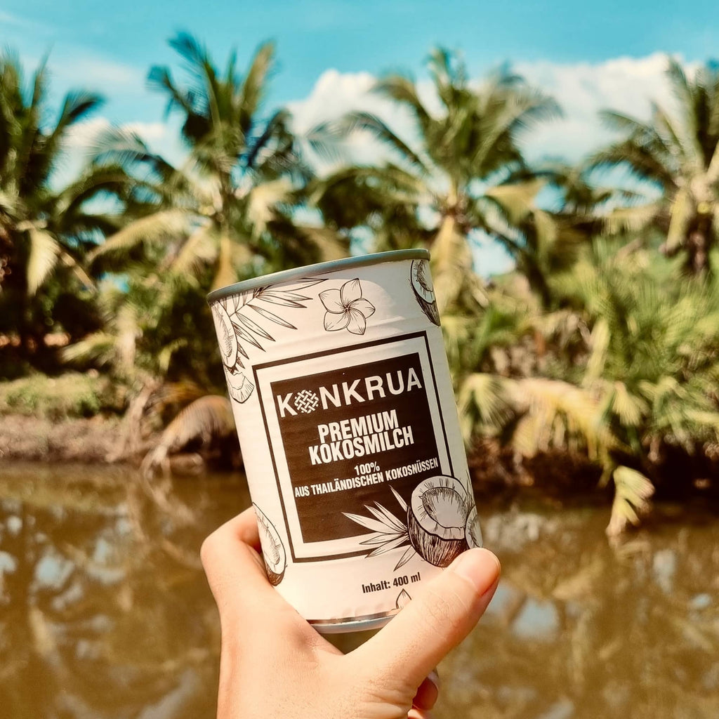 Eine Hand hält Konkrua Premium Kokosmilch vor der Kokosnussplantage. Im Hintergrund sieht man viele Kokospalmen und ein Kanal. KONKRUA Kokosmilch kommt direkt aus Thailand und schmeckt sehr cremig und aromatisch.